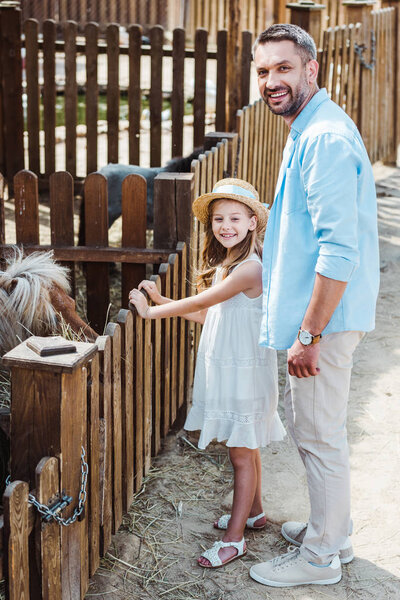 cheerful kid and man smiling while standing near pony in zoo 