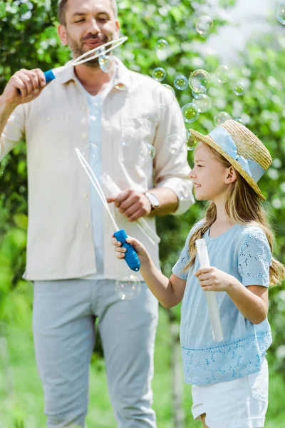 选择性焦点可爱的女儿在草帽拿着泡泡棒附近父亲吹肥皂泡 — 图库照片