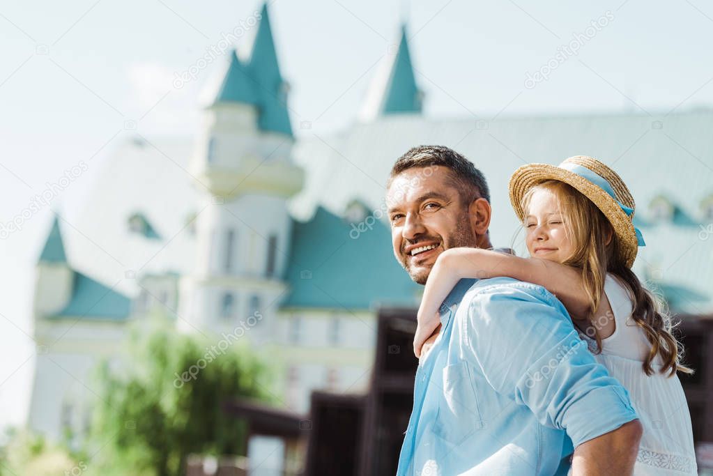 cheerful father piggybacking cute daughter in straw hat near castle 