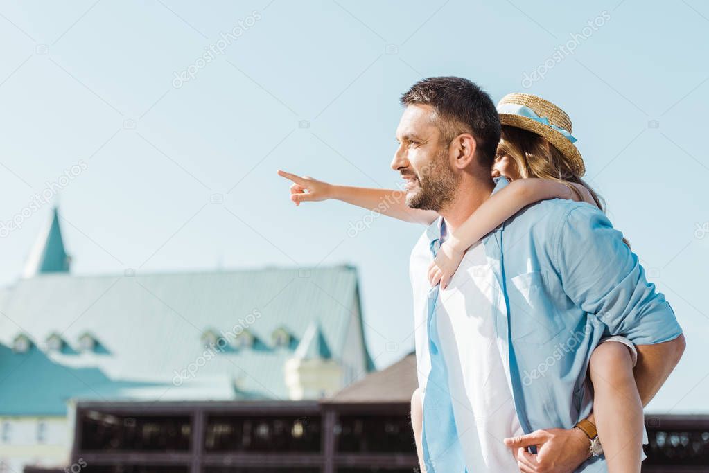 cheerful man piggybacking daughter pointing with finger near building 