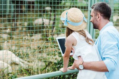 hayvanat bahçesinde dijital tablet tutan baba yakınında hasır şapkalı çocuk 