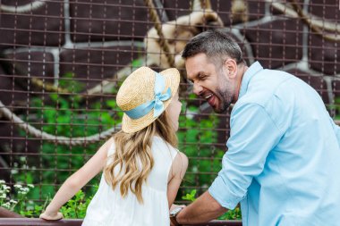 kid in straw hat looking at father showing tongue near cage with wild animal in zoo  clipart