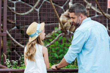 kid in straw hat looking at upset father near cage with wild animal in zoo  clipart