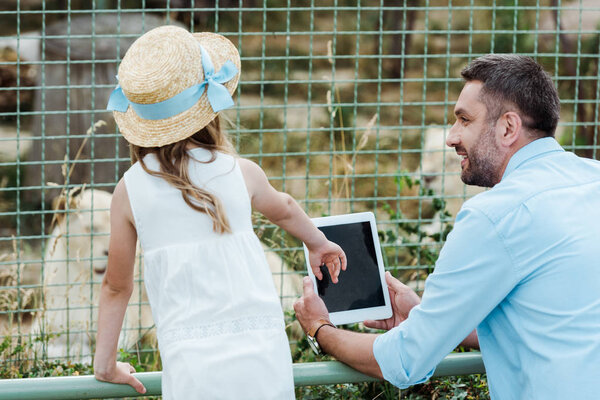 kid in straw hat gesturing near digital tablet with blank screen and father in zoo 