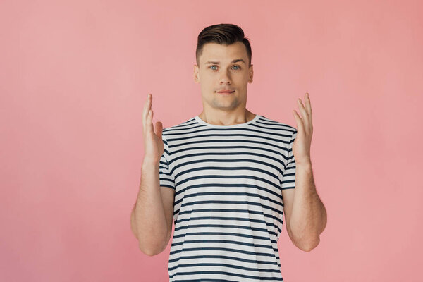 front view of handsome young man with hands up in striped t-shirt looking at camera isolated on pink