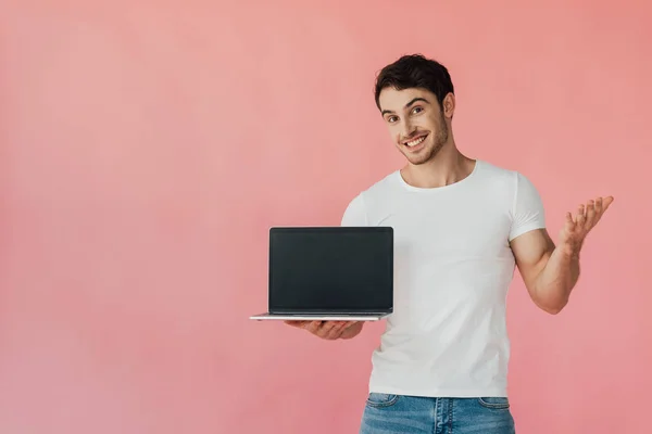 smiling muscular man in white t-shirt showing laptop with blank screen isolated on pink