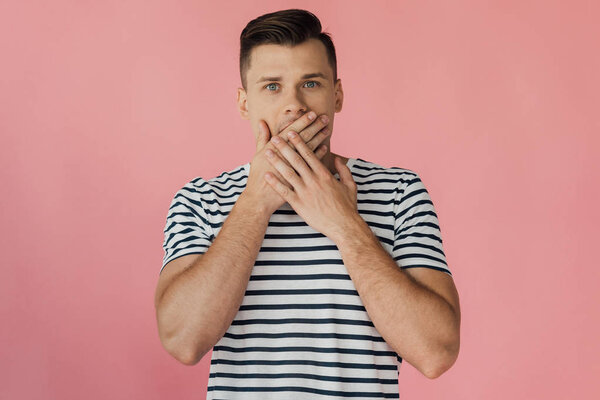 front view of shocked young man in striped t-shirt covering mouth with hands isolated on pink