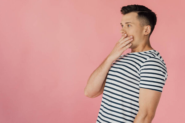 shocked young man in striped t-shirt covering mouth with hand isolated on pink