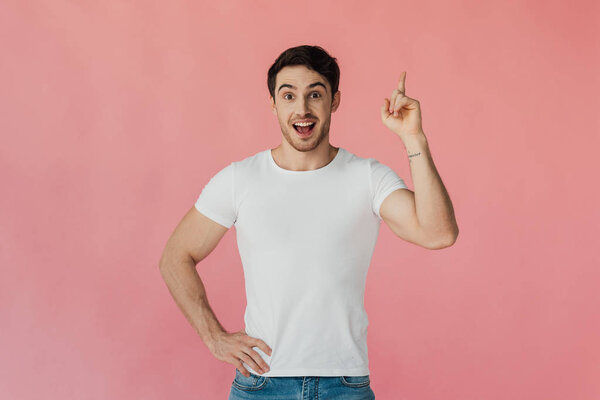 front view of excited muscular man in white t-shirt standing with hand on hip and showing idea sign isolated on pink