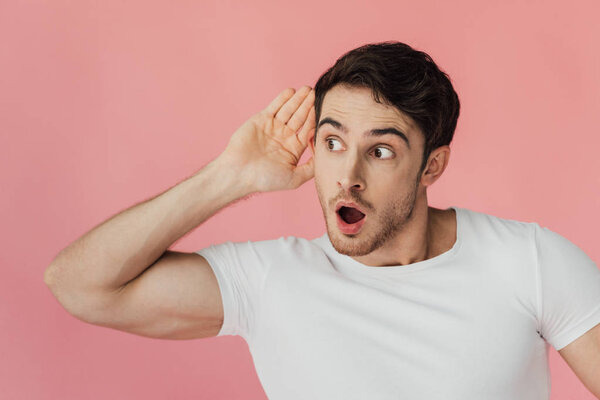 shocked muscular man in white t-shirt holding palm near ear and listening with open mouth isolated on pink