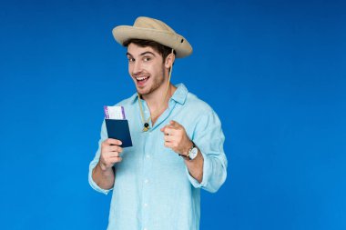 pasaport ve uçak bileti tutan safari şapka gülümseyen turist ve mavi izole kamera parmak ile işaret