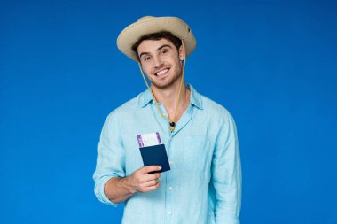 smiling traveler in safari hat holding passport and air ticket and looking at camera isolated on blue clipart
