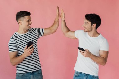 two smiling men using smartphones and looking at each other isolated on pink clipart