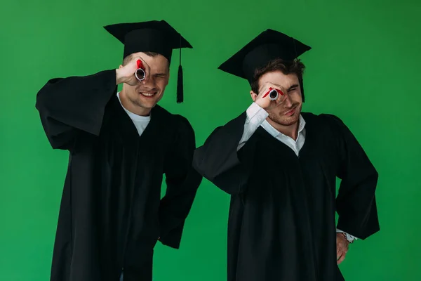 two happy students in academic caps holding diplomas isolated on green