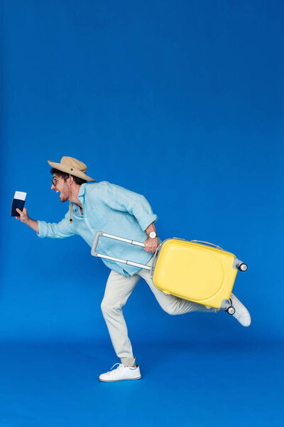 full length view of hurrying traveler in safari hat and sunglasses holding yellow suitcase and running on blue