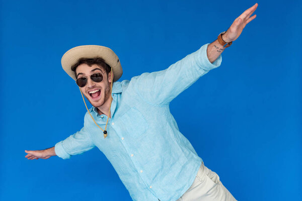excited tourist in safari hat and sunglasses laughing and waving hands isolated on blue