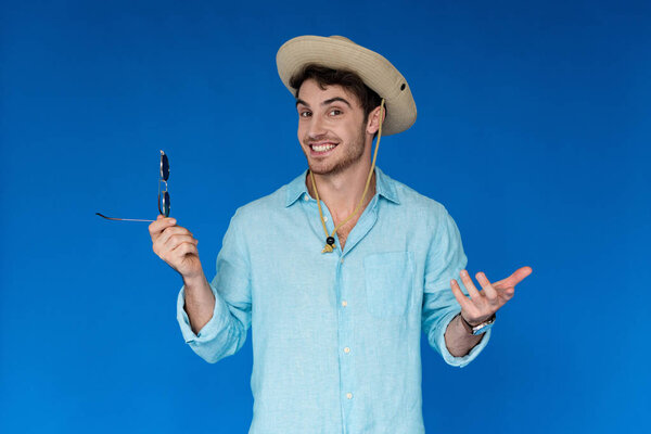 smiling traveler in safari hat holding sunglasses and looking at camera isolated on blue