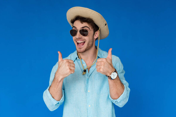 excited traveler in safari hat and sunglasses showing thumbs up isolated on blue