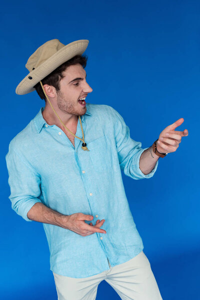 smiling traveler in safari hat gesturing and looking away isolated on blue