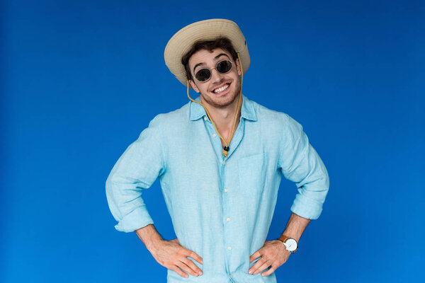 front view of smiling traveler in safari hat and sunglasses standing with hands on hips isolated on blue