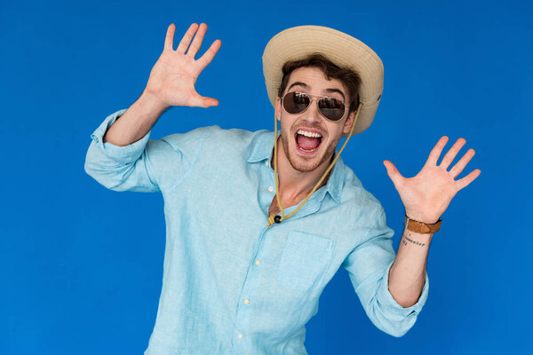 front view of excited traveler in safari hat and sunglasses standing with hands up isolated on blue