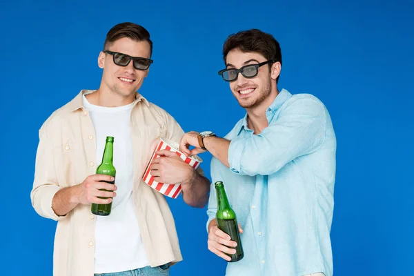 两个微笑的朋友在3D眼镜拿着啤酒和爆米花瓶隔离在蓝色 — 图库照片