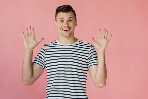 front view of excited smiling young man in striped t-shirt standing with hands up isolated on pink