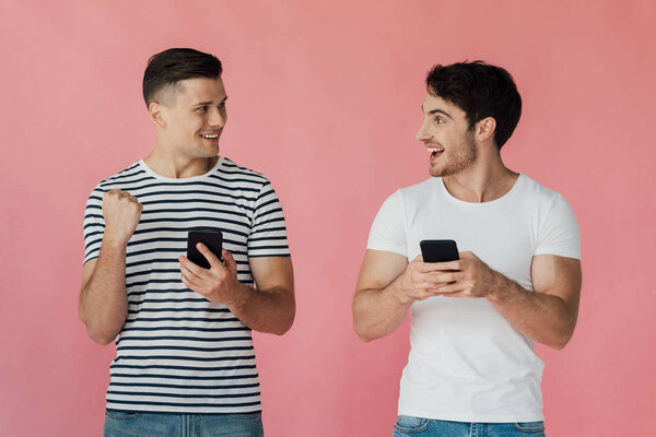 два улыбающихся мужчины с помощью смартфонов и глядя друг на друга изолированы на розовый
