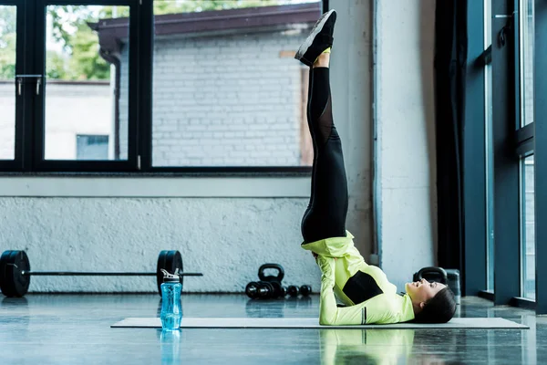 happy woman doing shoulder stand exercise on fitness mat near sport bottle