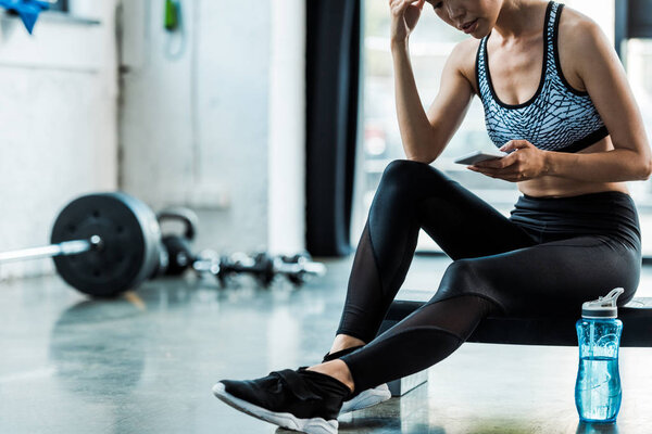 обрезанный вид спортсменки, сидящей на ступенчатой платформе и использующей смартфон
 