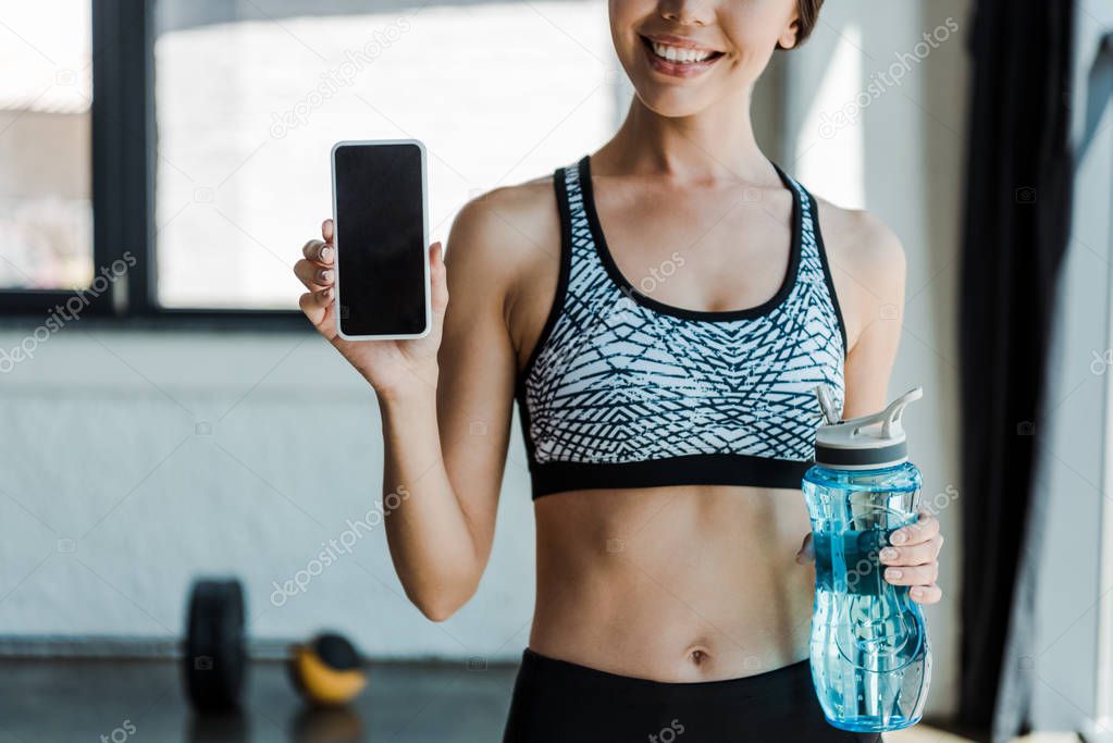 cropped view of cheerful girl holding sport bottle and smartphone with blank screen 