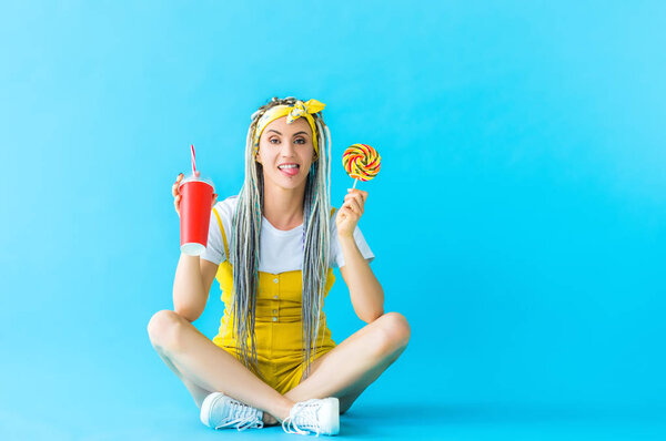 girl with dreadlocks and soda drink sticking out tongue and holding lollipop on turquoise