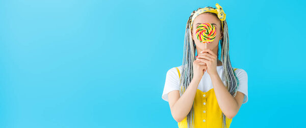 panoramic shot of girl with dreadlocks covering face with lollipop isolated on turquoise