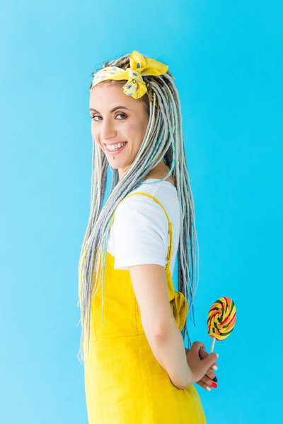 smiling girl with dreadlocks looking at camera while holding lollipop isolated on turquoise