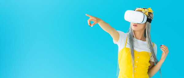 panoramic shot of girl with dreadlocks in virtual reality headset pointing with finger isolated on turquoise