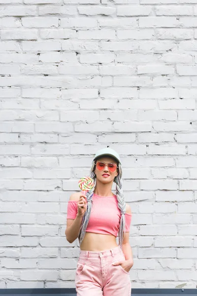レンガの壁の近くにロリポップを保持している帽子とサングラスの女の子 — ストック写真