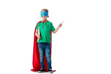 full length view of preschooler kid in mask and hero cloak holding digital tablet and pointing with finger on white clipart