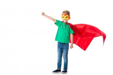 full length view of smiling preschooler boy in mask and red hero cloack standing with fist up isolated on white clipart