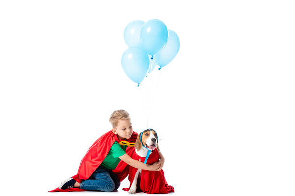 Дитя-дошкольник в красном плаще героя обнимает собаку возле голубых воздушных шаров партии, изолированных на белом
