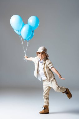 bir bacak üzerinde ayakta gözlüklü Explorer çocuk tam uzunlukta görünümü ve gri üzerinde mavi balonlar tutarak