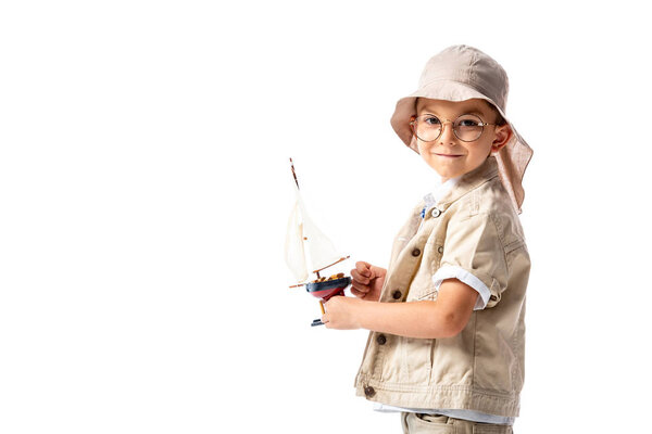 smiling explorer child in glasses and hat holding toy ship isolated on white