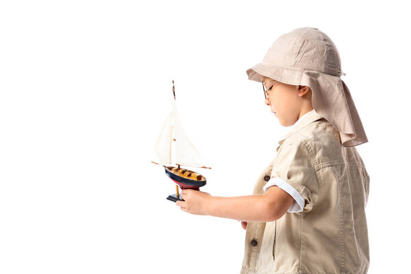 side view of explorer child in glasses and hat holding toy ship isolated on white
