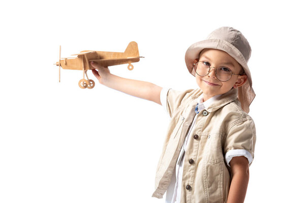 smiling explorer boy in glasses and hat holding wooden toy plane isolated on white