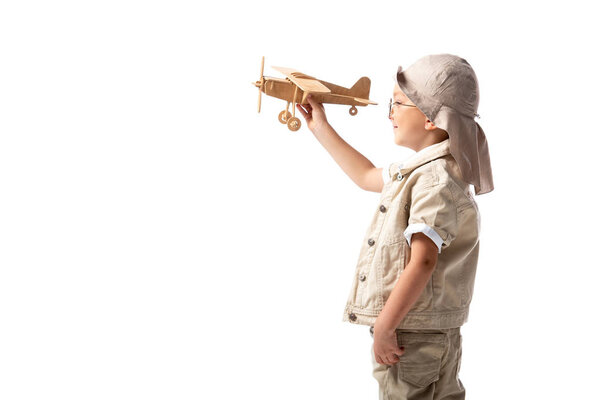 side view of smiling explorer boy in glasses and hat holding wooden toy plane isolated on white