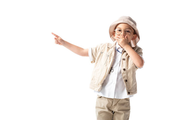 shocked explorer boy in hat and glasses covering mouth with hand and pointing with finger isolated on white