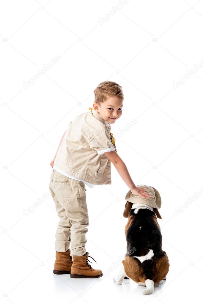 full length view of preschooler explorer boy stroking beagle dog in hat and smiling on white