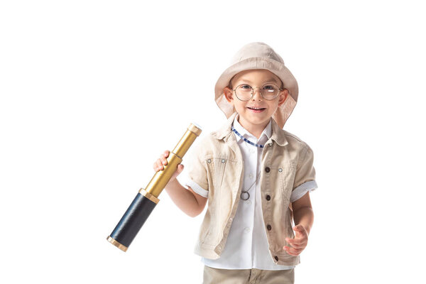 front view of smiling explorer boy in glasses and hat holding spyglass isolated on white