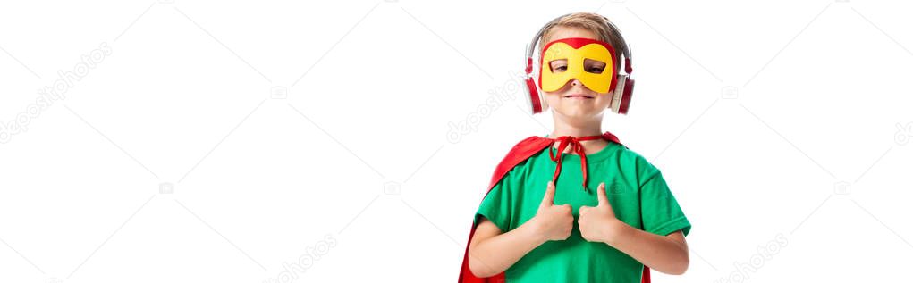 panoramic shot of smiling boy in hero costume listening music in headphones and showing thumbs up isolated on white