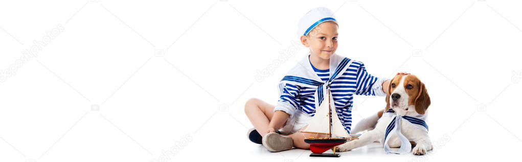 panoramic shot of smiling boy in sailor suit with toy ship stroking beagle dog on white
