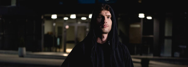 панорамный снимок вора в черной толстовке, смотрящего на камеру на парковке ночью
